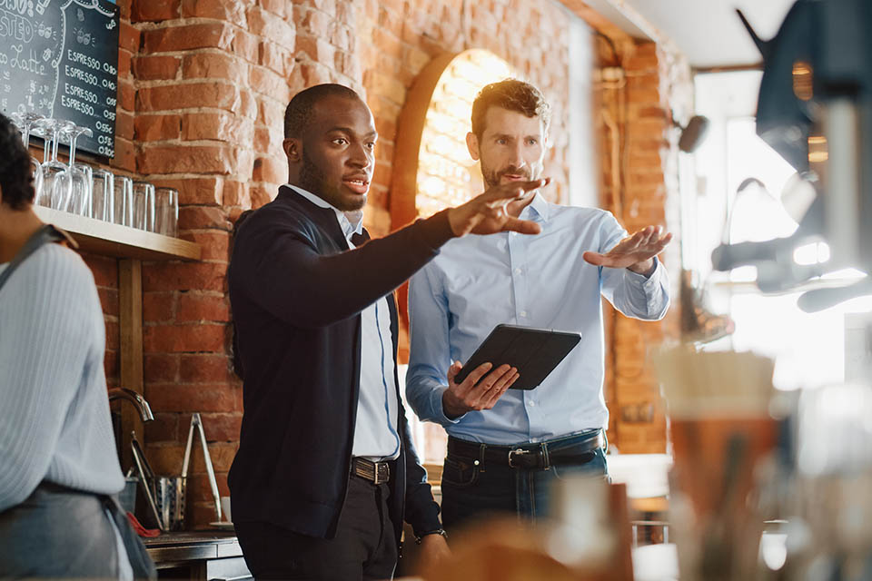 Multicultural Coffee Shop Owners Meeting Behind the Counter and Working on Tablet Computer and Checking Inventory in a Cozy Loft-Style Cafe. Successful Restaurant Managers and Barista at Work.