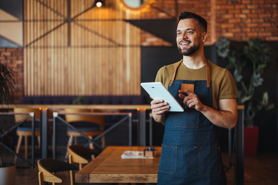 A cheerful male waiter in a stylish apron stands with a digital tablet, ready to take orders in a contemporary restaurant setting.
